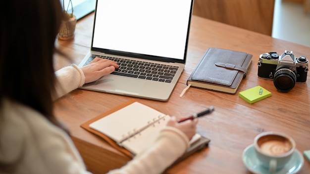 Young woman blogger freelance photographer working on laptop while taking some notes on diary