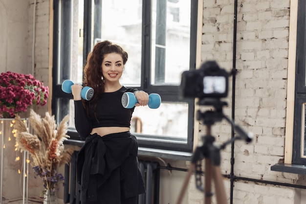 Young woman blogger with camera on tripod recording fitness blog and trains with dumbbells Fitness sport and video blogging concept