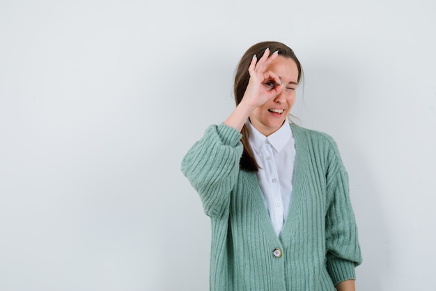 Young woman in blouse, cardigan showing ok sign on eye and looking curious , front view.