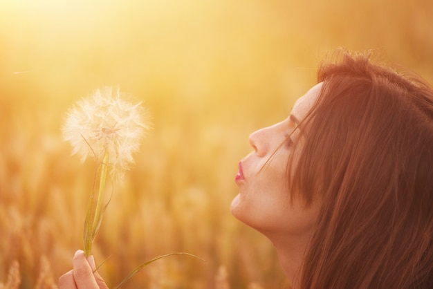 Young woman blowing dandelion in autumn landscape at sunset