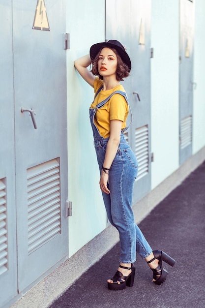 Young woman in blue denim overalls and yellow tshirt with black hat sensual looking at camera while posing near industrial building . outdoor shot in the summertime