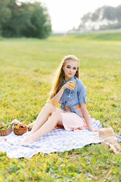 Young woman in a blue denim shirt and pink skirt in the garden at a picnic holding a glass of juice in her hand