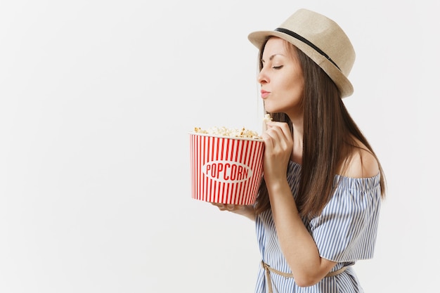 Young woman in blue dress, hat watching movie film holding eating popcorn from bucket isolated on white background. People, sincere emotions in cinema, lifestyle concept. Advertising area. Copy space.