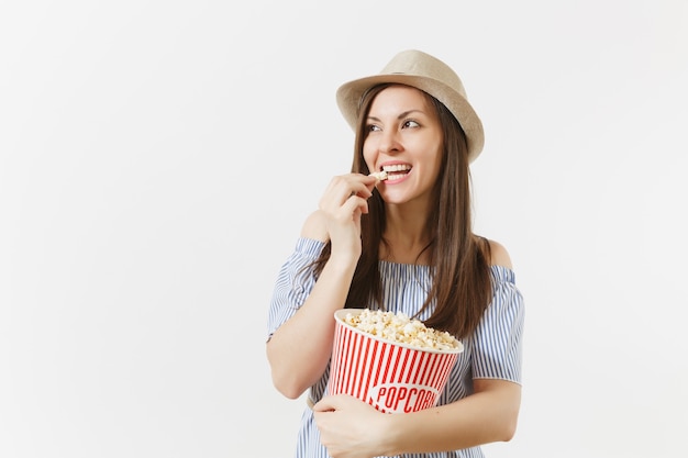 Young woman in blue dress, hat watching movie film holding eating popcorn from bucket isolated on white background. People, sincere emotions in cinema, lifestyle concept. Advertising area. Copy space.