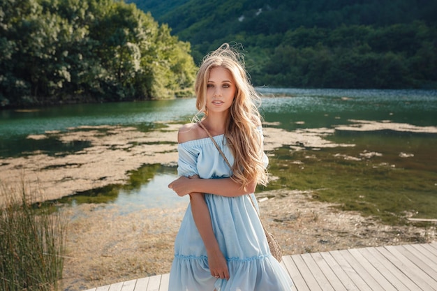 Young woman in blue dress standing alone on footbridge and staring at lake Mist over water Chilly morning Beautiful fashionable girl with a straw bag posing on a mountain lake