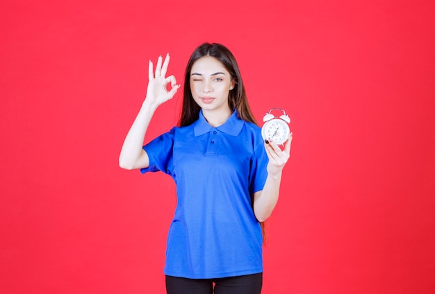 Young woman in blue shirt holding an alarm clock and showing positive hand sign