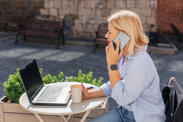 Young woman in a blue shirt is working on a laptop in a cafe and talking on the phone