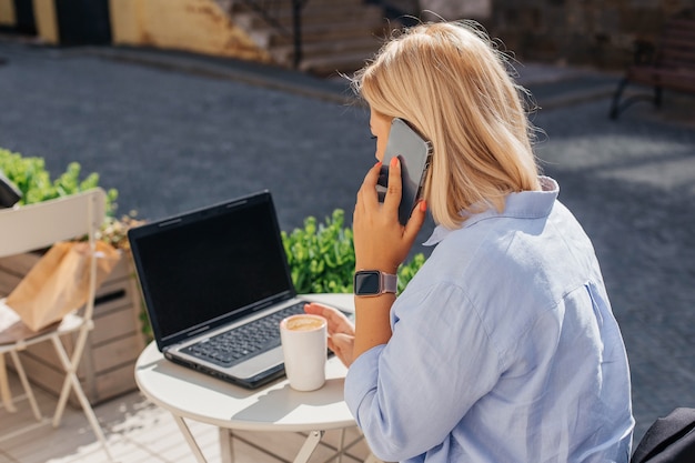 Young woman in a blue shirt is working on a laptop in a cafe and talking on the phone