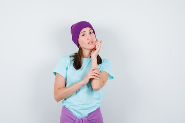 Young woman in blue t-shirt, purple beanie leaning chin on hand, with hand on arm while posing and looking charming , front view.