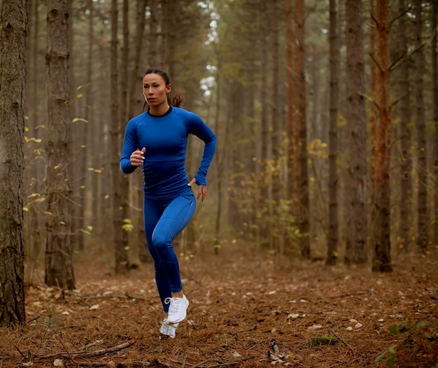 Young woman in blue track suit running toward camera on the forest trail at autumn