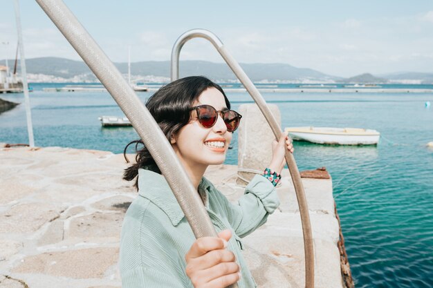 Young woman in a boat dock smiling during a sunny day while using sunglasses beach day