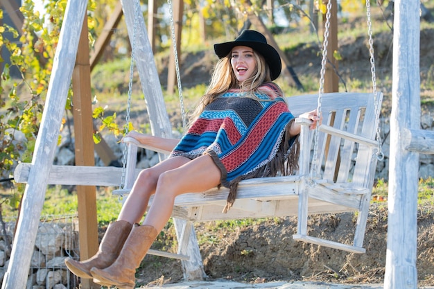 Young woman in boho style swinging on a swing in park Beautiful hipster woman relaxing at a swing in garden Summer fashion portrait of sensual girl in hat Outdoor portrait of a smiling happy girl