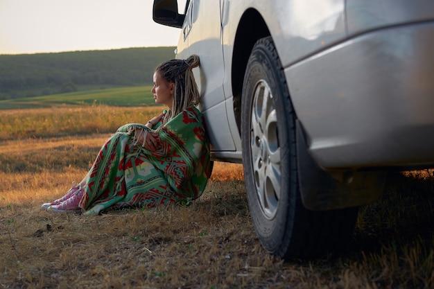 A young woman in a bright green blanket sits leaning against a car against the background of a green...