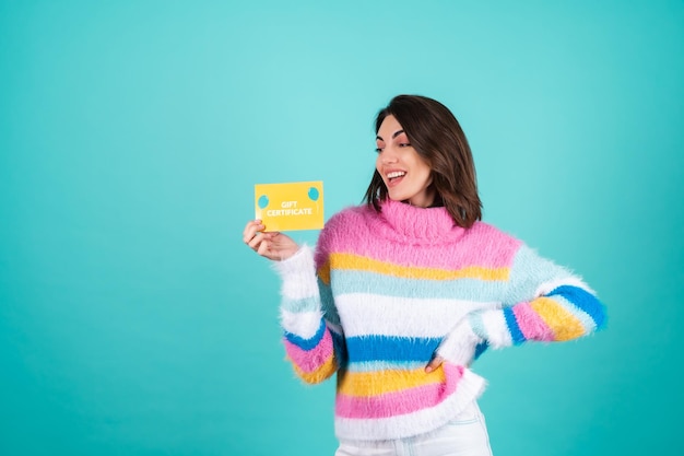 Young woman in a bright multicolored sweater on blue holds a gift certificate