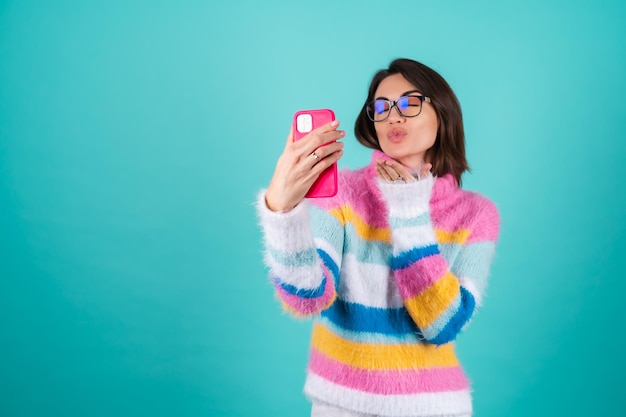 A young woman in a bright multicolored sweater on blue with glasses, holds a phone, takes a selfie