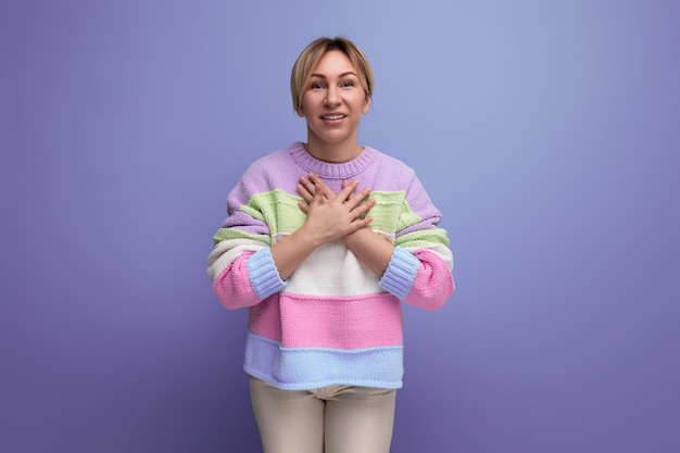 Young woman in a bright sweater gives thanks holding her hands on a heart on a purple background