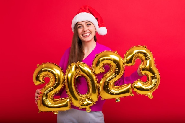 A young woman in a bright sweater wearing a Santa Claus hat The brunette holds the figures of the new year 2023