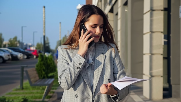 Young woman in a business suit talking on the phone and looking at documents walking down the street