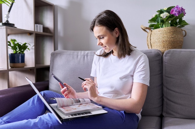 Young woman buys goods, cosmetics, clothes online. In the hands of paper catalog laptop smartphone, female makes purchases at home, sitting on the couch