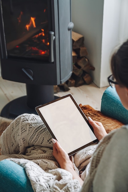 Young woman by the fireplace, sitting in a cozy armchair, with a warm blanket, using a tablet