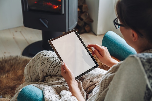 Young woman by the fireplace, sitting in a cozy armchair, with a warm blanket, using a tablet