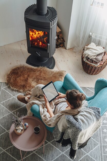 Young woman by the fireplace, sitting in a cozy armchair, with a warm blanket, using a tablet