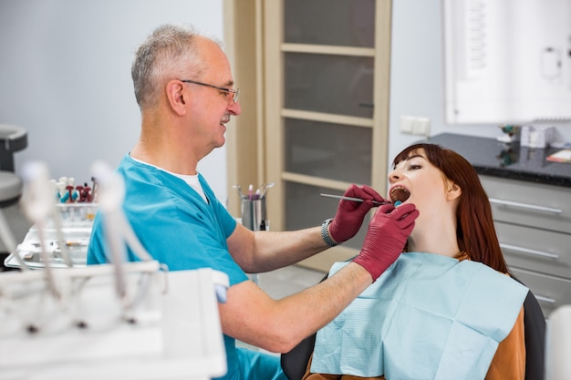 Young woman came to see the dentist, sitting in the dental chair with open mouth. Senior smiling man dentist checking her teeth. Happy patient and dentist concept.