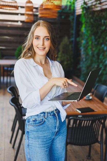 Young woman in casual clothes drinking coffee and talking on the phone working on a green terrace at a laptop looking at the screen and working on a project remotely from work in a cafe