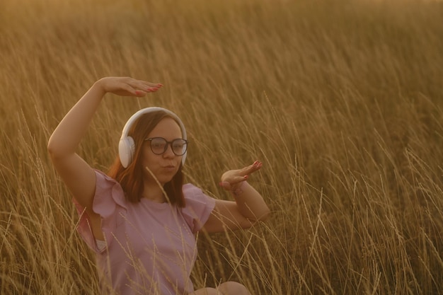 A young woman in casual clothes and glasses listens to music with white headphones and dancing sitting in the dry grass