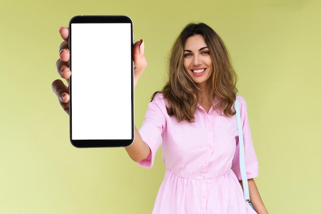 Young woman in casual wear isolated on green background holds a phone with a blank white screen