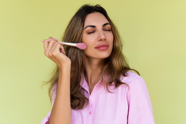 Young woman in casual wear isolated on green background playful positive with blush brush natural daily makeup