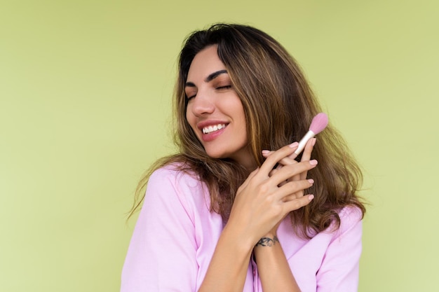 Young woman in casual wear isolated on green background playful positive with blush brush natural daily makeup