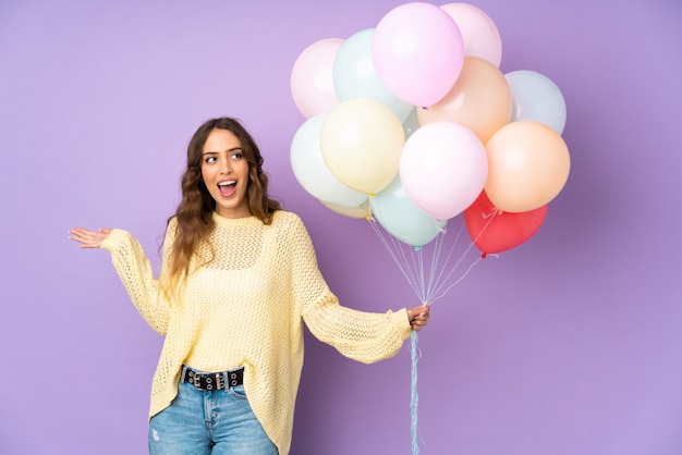 Young woman catching many balloons over on purple wall with surprised facial expression