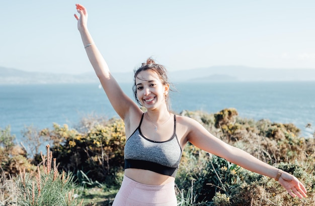 Young woman celebrates finishing a outdoor exercise session on the coastline Dancing and raising arms to the air Happy young people training during a sunny summer day preparing for the beach body