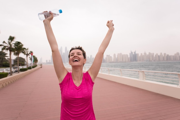 young woman celebrating a successful training run on the promenade by the sea with a bottle of water and her hands raised in the air with a big city in the background