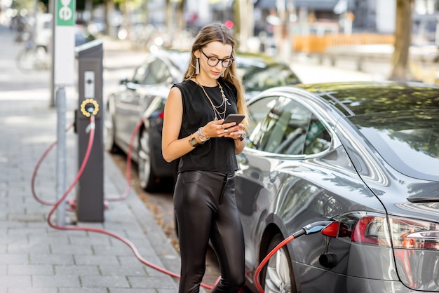 Young woman charging electric car standing with smart phone outdoors on the street in Rotterdam city