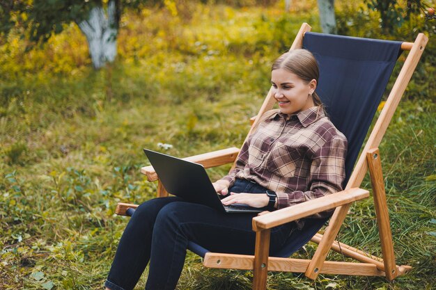 Young woman in a checkered shirt sitting in a chair outdoors in a garden and working on a laptop remote work female freelancer