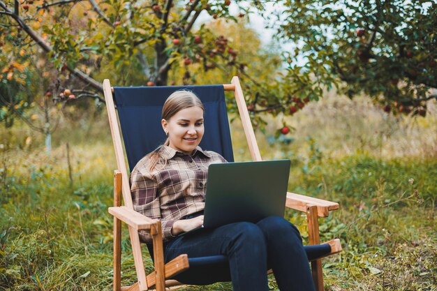 Young woman in a checkered shirt sitting in a chair outdoors in a garden and working on a laptop remote work female freelancer