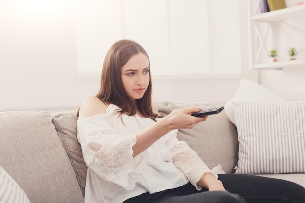 Young woman choosing channels with TV remote while sitting on beige couch at home, copy space