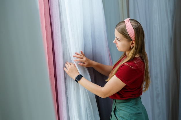 Photo young woman choosing fabric for new curtains in a store. samples of the curtain hang on hangers on a rail in the store. samples of textures of fabric, tulle and furniture upholstery.