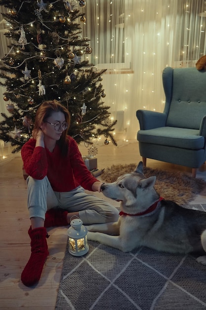 Young woman on Christmas evening together with her pets petting a dog while sitting on the floor next to the Christmas tree