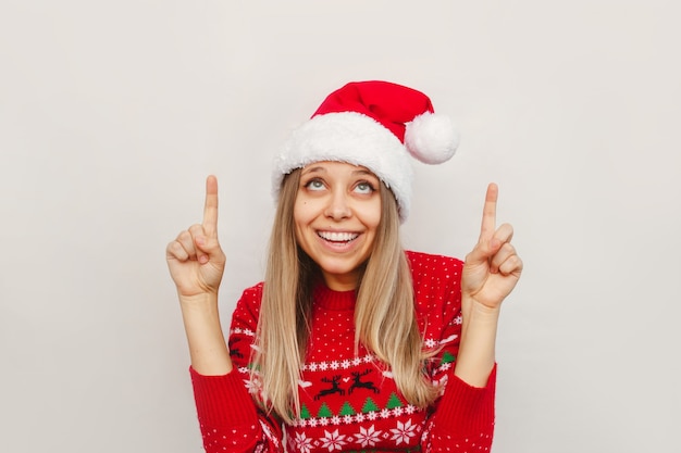 A young woman in a Christmas red sweater Santa hat points up at empty copy space for text or design