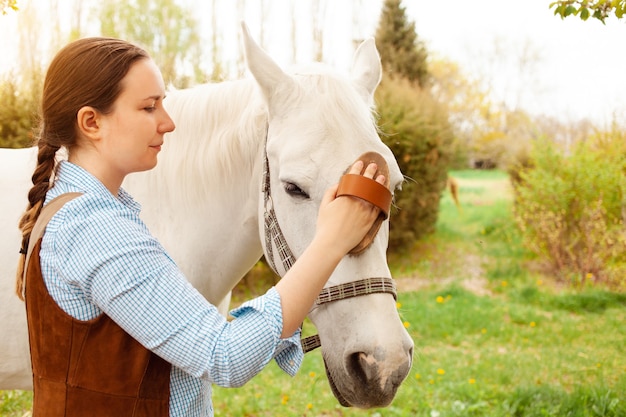 A young woman cleans a white horse with a yellow brush in nature