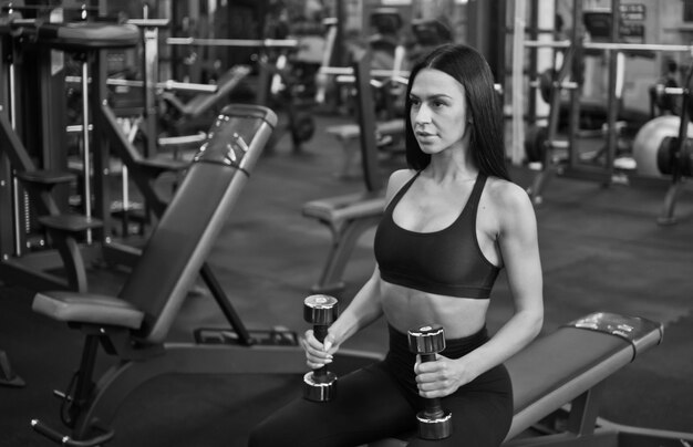 Young woman concentrated on training holds dumbbells in hands while sitting on a bench in gym