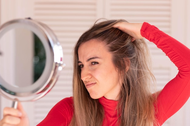 Young woman concern about her hair loss in autumn time while looking to round hand mirror in red outfit