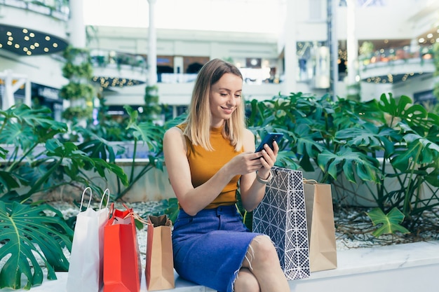 Young woman consumer in the mall browses chat and uses using a smartphone. female standing with a mobile phone in her hands in shopping center. indoor. happy shopper girl with gift bags make purchases