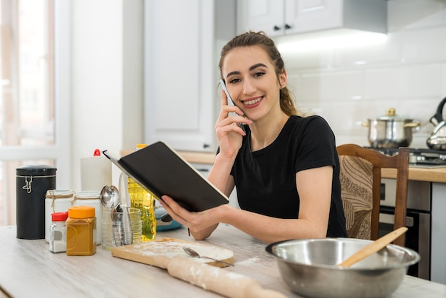 Young woman cooking cake with flour reading notepad recipe in kitchen table