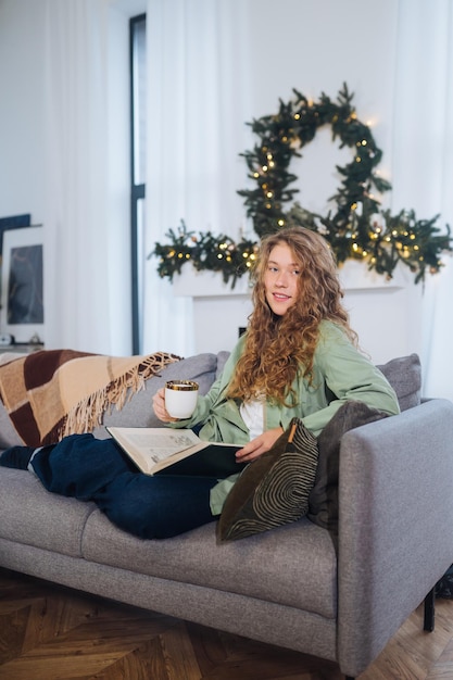 Young woman on the couch reads a book and enjoys coffee