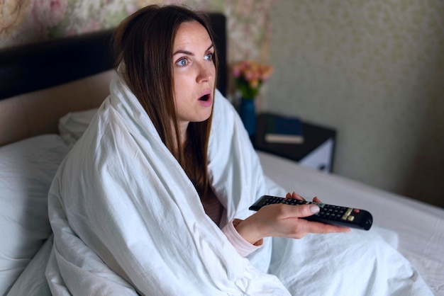 Young woman covered blanket relaxing in bed in the evening and watching tv