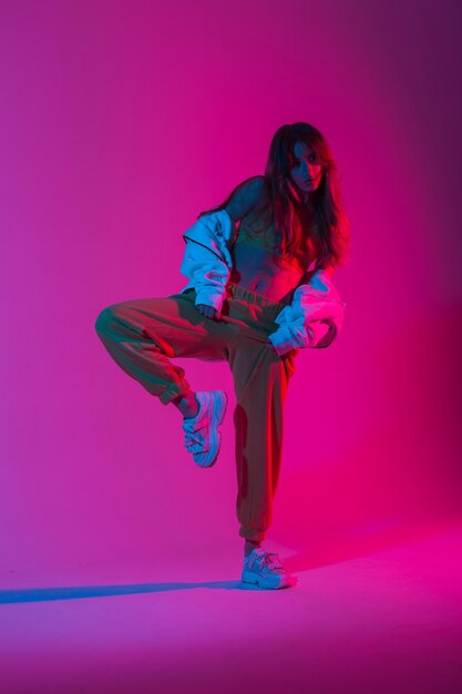 Young woman dancer in a stylish top in trendy pants in sneakers posing on one leg in a room with bright pink ultraviolet light. Cool modern stylish girl dancing indoors with neon multi-colored color.